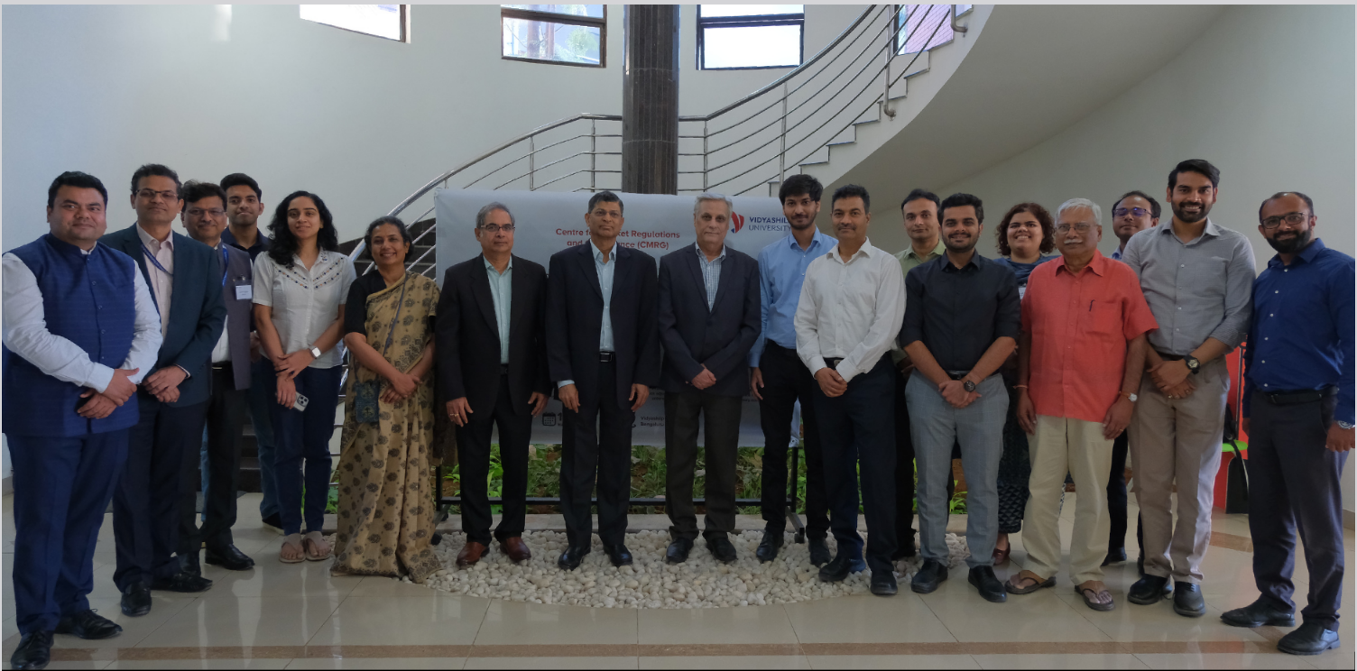 A medium shot in natural light of a group of eighteen multi-generational professionals, fifteen men and three women, with various skin tones, posing together in the brightly lit modern atrium of Vidyashilp University with natural white tiled flooring and a white pebbled border on the floor in front of a curved steel staircase and background. All attendees look directly at the camera with broad smiles and serious expressions as they stand with hands clasped, at their side or in their pockets, in front of a banner that is about the the Certificate Course on Enforcement Process for Regulators, Regulated Entities, and Regulatory Practitioners held on May 10-11, 2025.